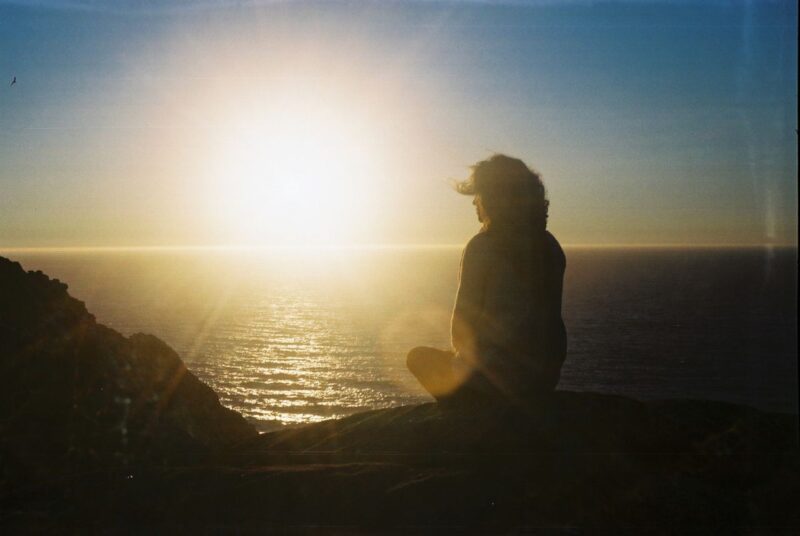 Silhouette of a person meditating on a cliff during sunset by the ocean, embodying tranquility.